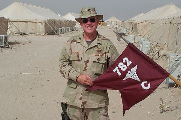 A soldier in desert camouflage holding a flag with tents in the background.